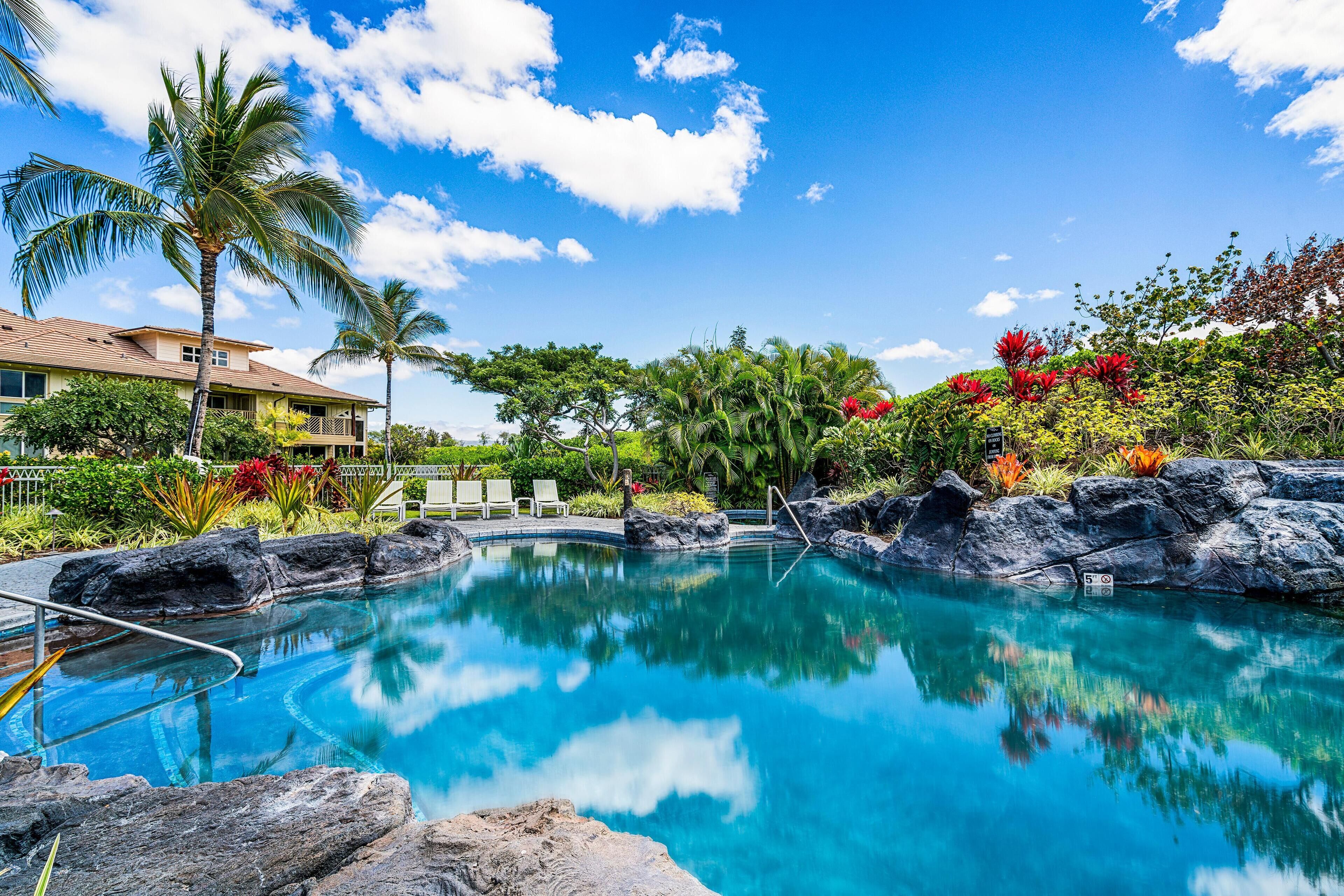 A clear blue pool surrounded by rocks and palm trees, with a building in the background.