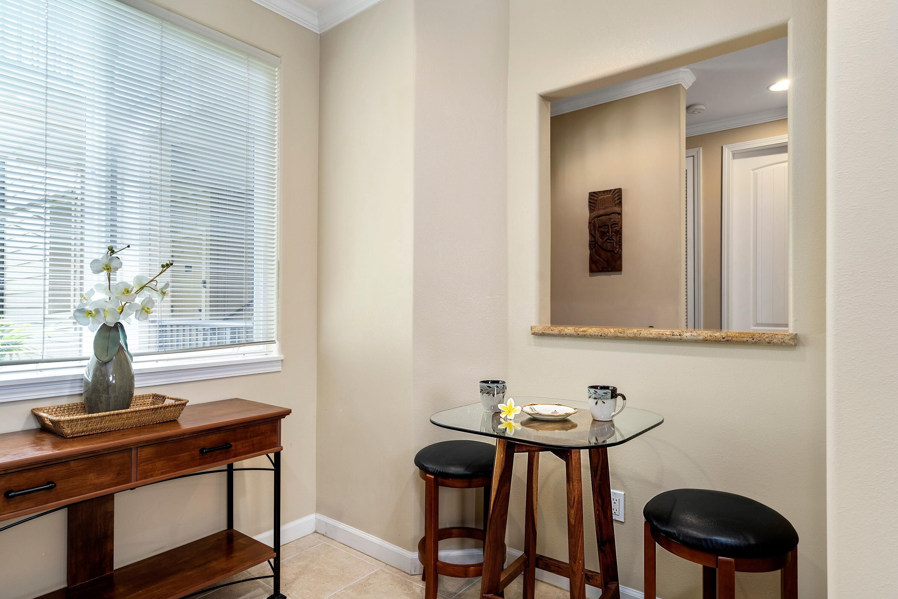 A corner room with a dining table, two bar stools, a vase with flowers, and a window with blinds.