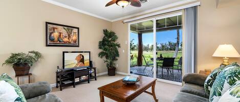 A living room with a ceiling fan, a television, a coffee table, and a large window with a view of the outdoors.
