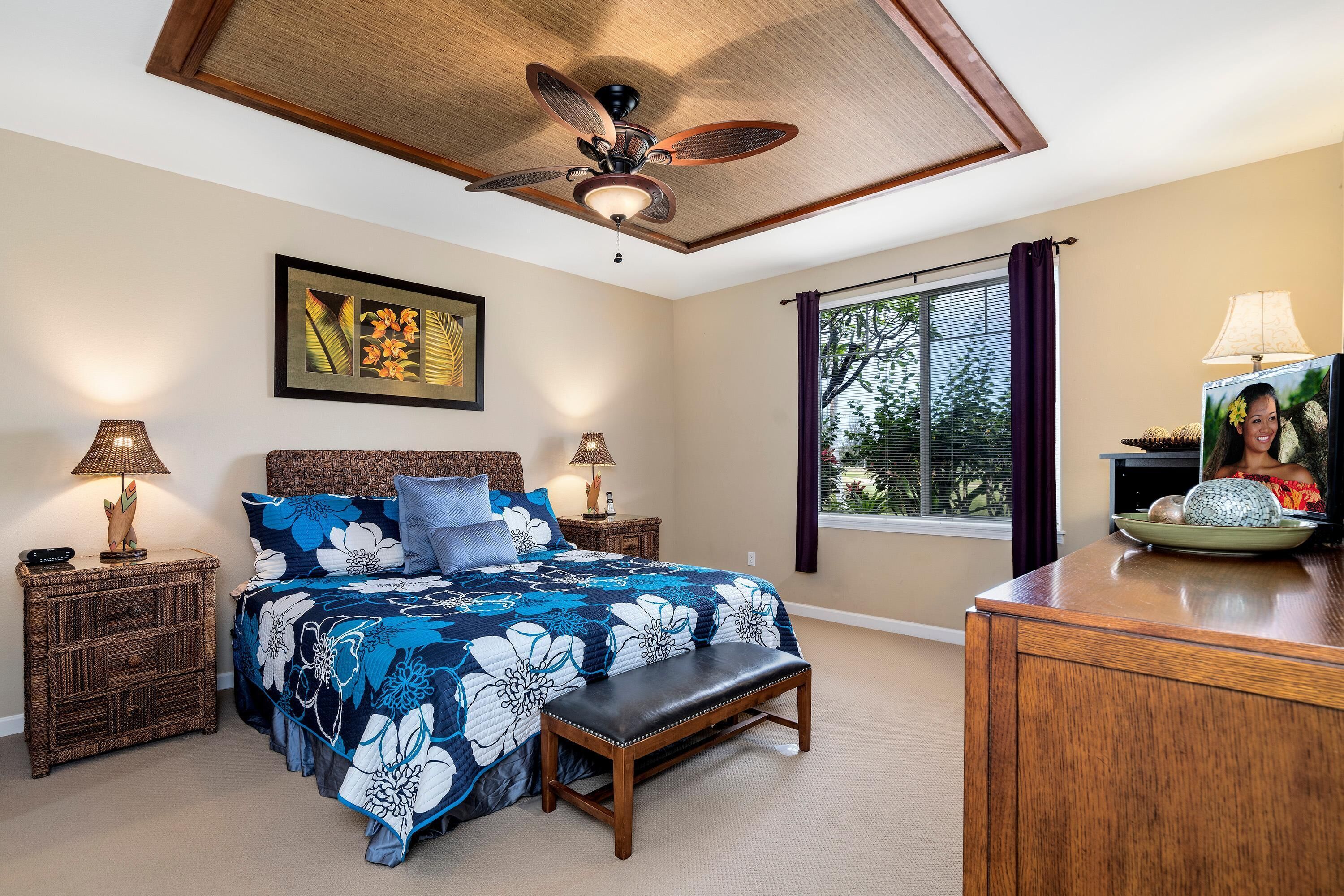 A bedroom with a bed, two nightstands, a bench, a ceiling fan, a window with curtains, and a desk with a lamp.