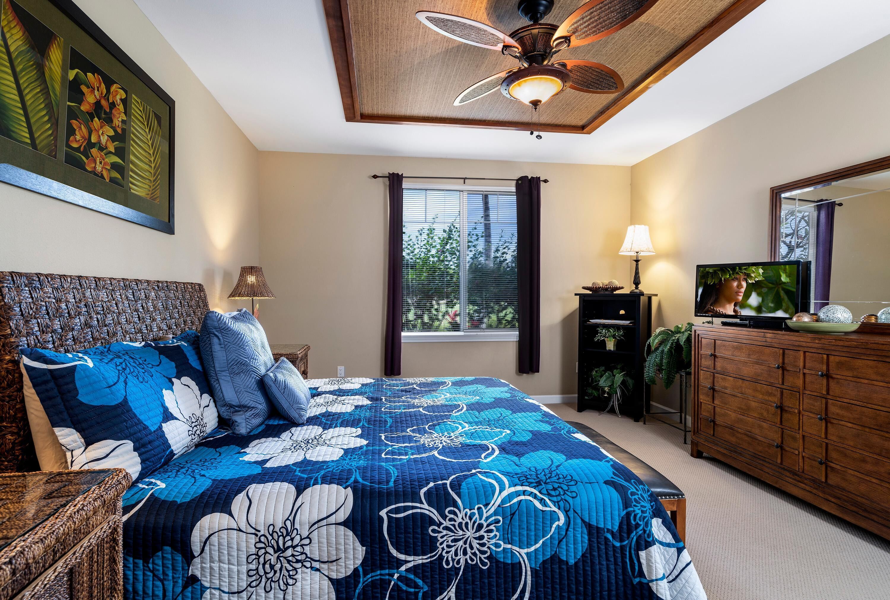 A bedroom with a large bed, a ceiling fan, a window with curtains, a dresser, and a television.