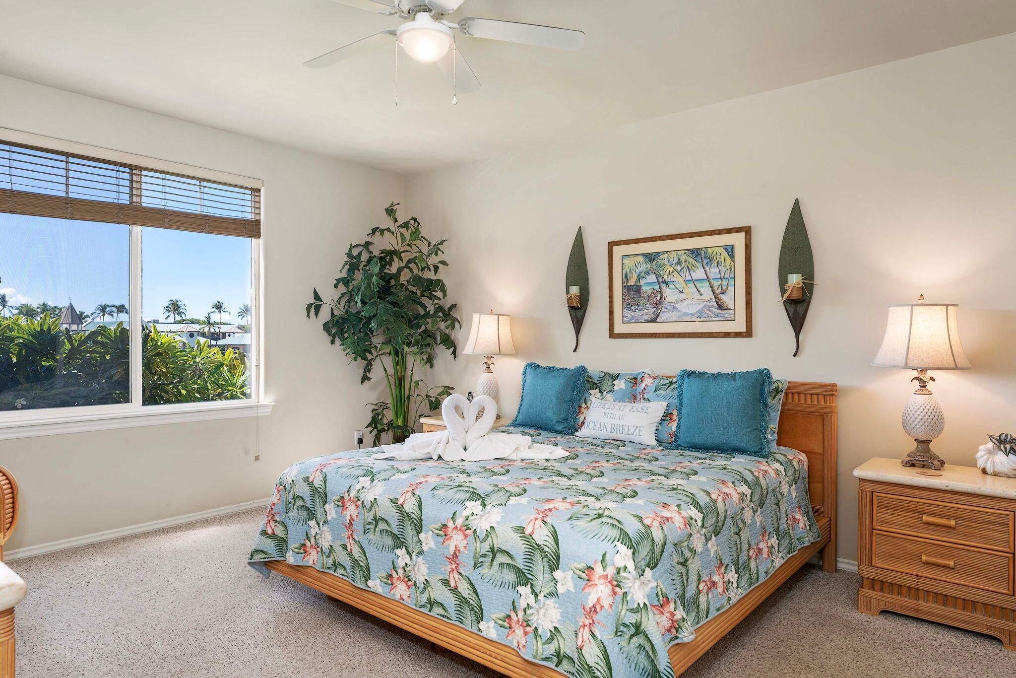 A bedroom with a large bed, two bedside lamps, a nightstand, a potted plant, and a window with a view of palm trees.