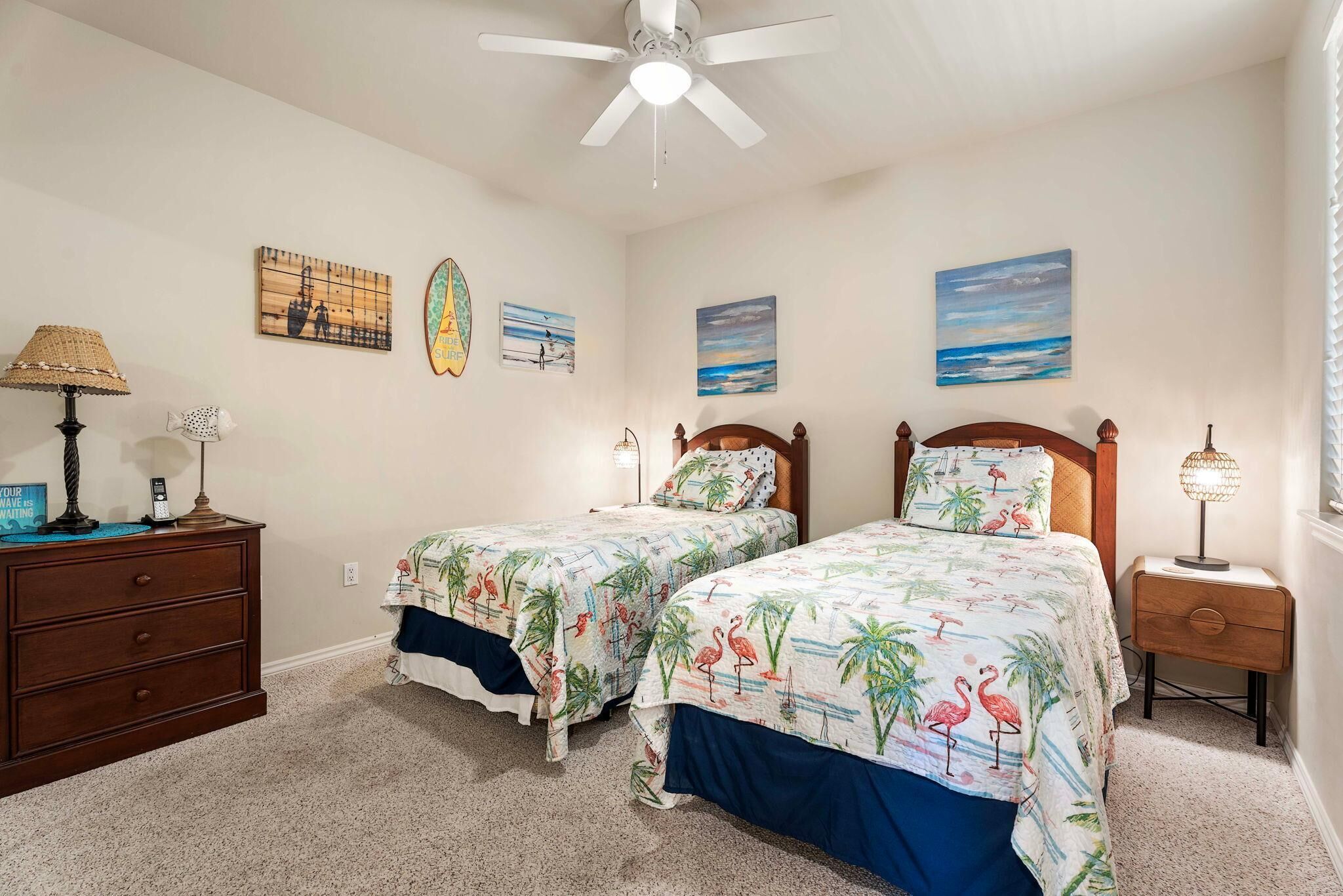 A bedroom with two beds, a ceiling fan, a nightstand, and wall art.