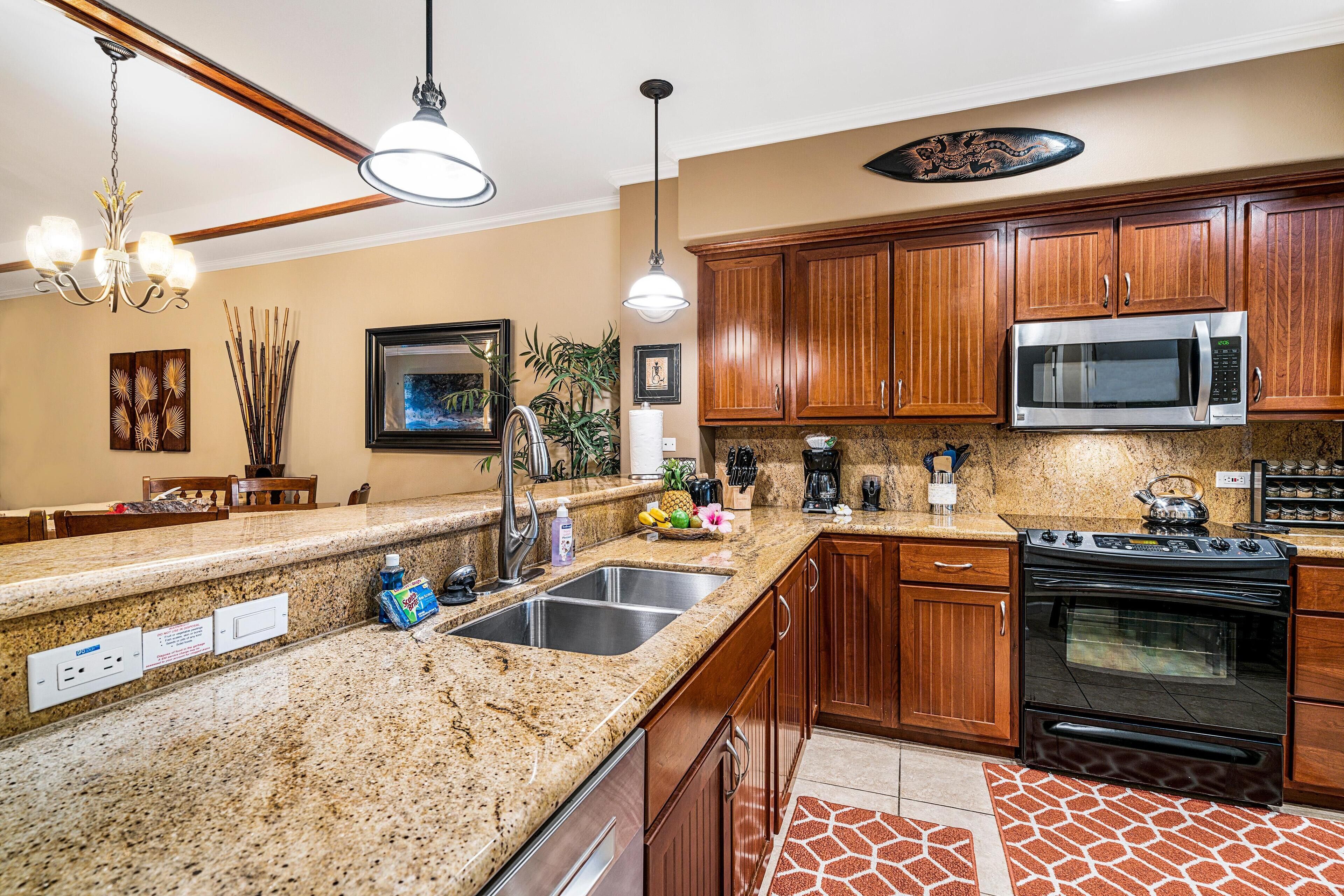 A kitchen with granite countertops, wooden cabinets, a stainless steel sink, a microwave, a stove, and pendant lighting.