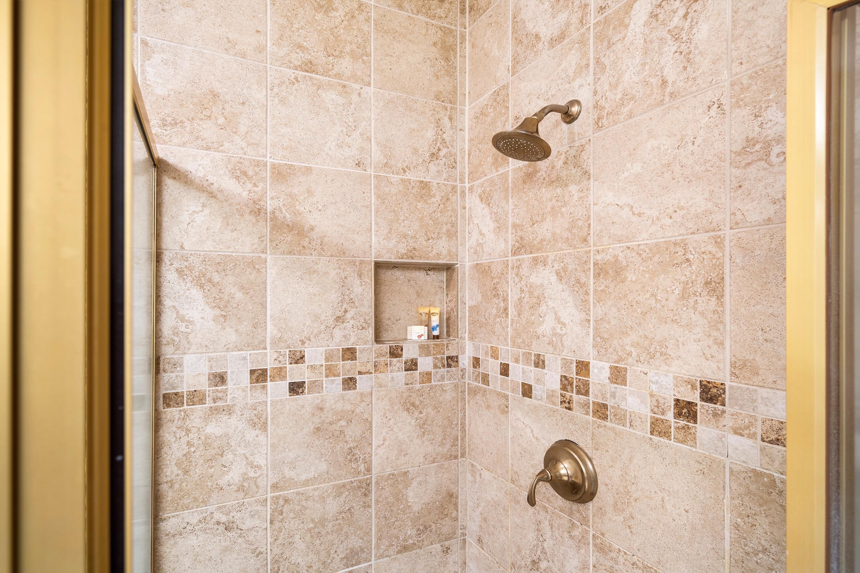 A shower area with beige tiles, a showerhead, and a small shelf with toiletries.