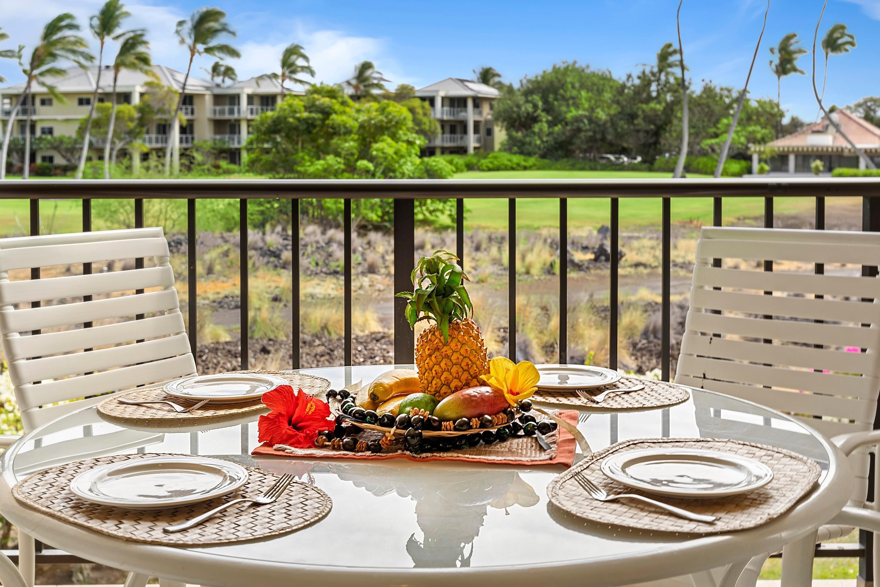 A dining table set for four with a tropical fruit centerpiece, white chairs, and a view of a golf course and buildings in the background.