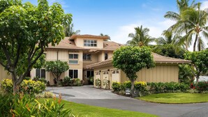 A two-story building with a beige facade, a curved driveway, and lush landscaping including palm trees and flowering plants. - Waikoloa Beach Villas C3 (Waikoloa)