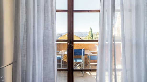 An open door leading to a balcony with a table and chairs, a view of mountains, and a clear sky. - Il Borgo apartments - B5 (Finale Ligure)