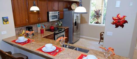 A modern kitchen with granite countertops, wooden cabinets, stainless steel appliances, and a breakfast bar with red placemats.