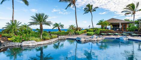 A pool area with palm trees, a lounge with umbrellas, and a view of the ocean.