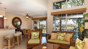 A spacious living area with wicker furniture, a glass coffee table, and large windows offering a view of the landscape. - Golf Villas at Mauna Lani - Unit K5 (Kamuela)
