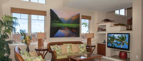 A spacious living room with a large window, a wicker sofa, a flat-screen TV, and a bookshelf.