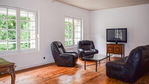 A spacious living room with hardwood flooring, two leather armchairs, a glass coffee table, and a television on a wooden cabinet. Large windows provide a view of greenery outside. - 3 bedroom stunning home in Langoat (Langoat)