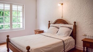 A bedroom with a wooden bed, white bedding, a nightstand, a wall-mounted light, and a window with a view of greenery. - 3 bedroom stunning home in Langoat (Langoat)