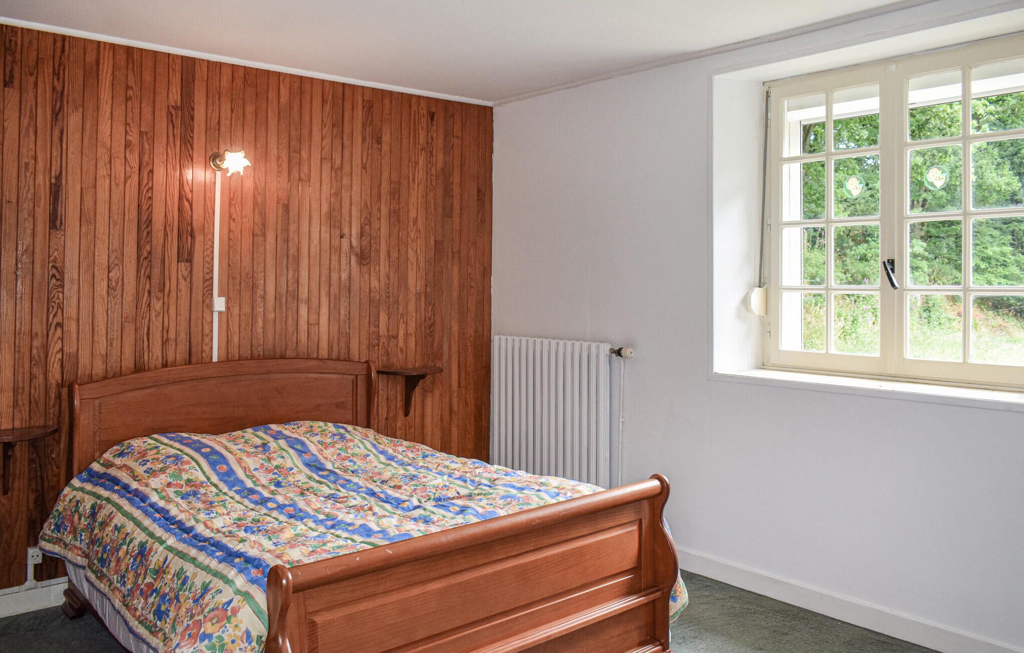 Un dormitorio con cabecera de madera, un radiador y una ventana con vista a la vegetación.