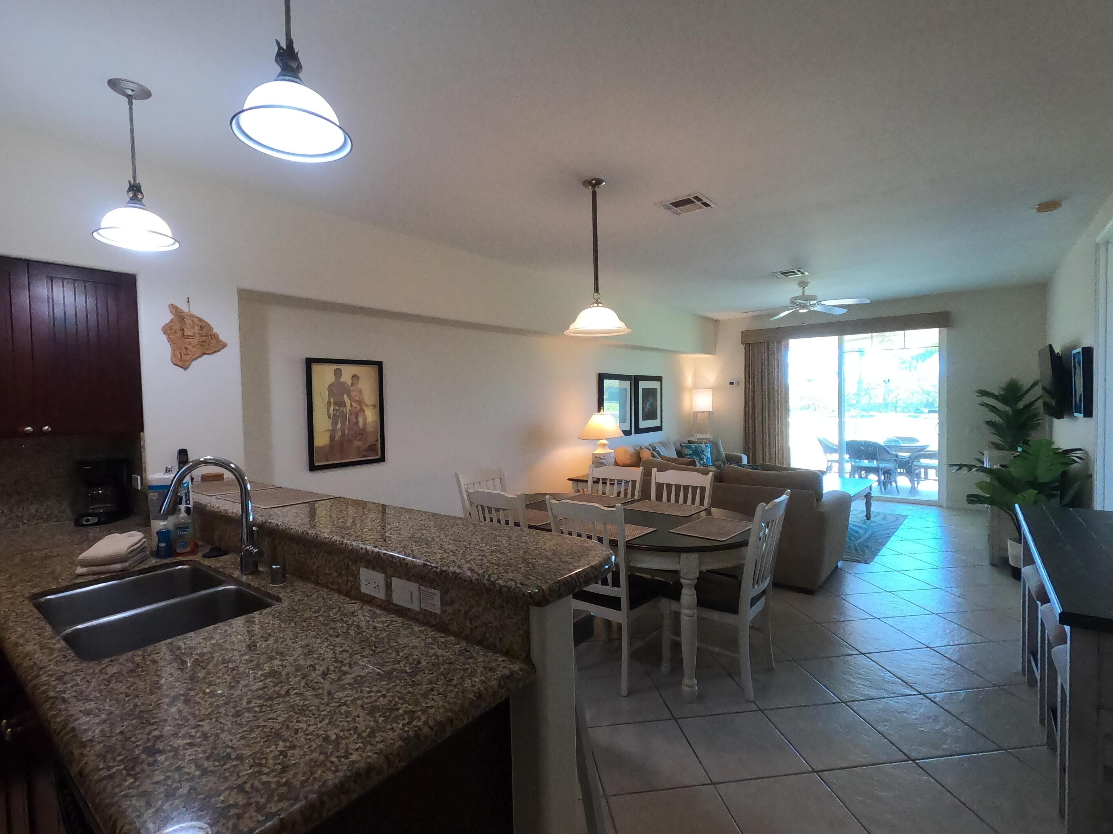 A kitchen with granite countertops, a dining area with chairs and a table, a living room with a sofa, and a large window.