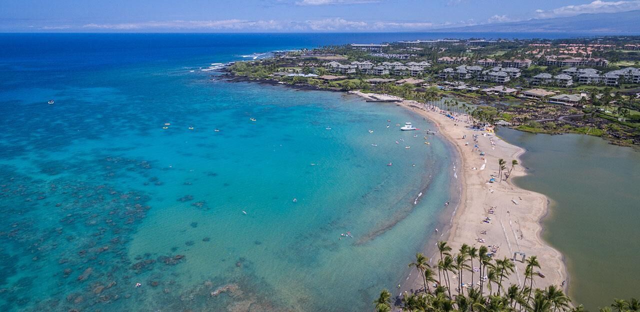 Vue aérienne d’une zone côtière avec une plage, des eaux turquoise et limpides, et une zone résidentielle avec des maisons.