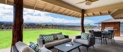 An outdoor patio with wicker furniture, a ceiling fan, and a view of a grassy area and distant mountains.
