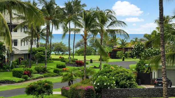 A residential area with a well-maintained lawn, palm trees, and a view of the ocean. - Vista Waikoloa B206 (Waikoloa)