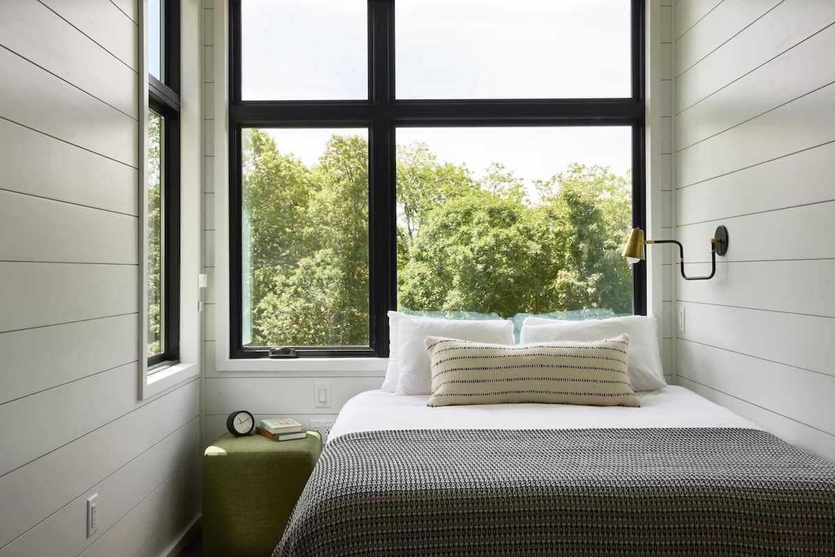 Un dormitorio con un ventanal grande, una cama con ropa de cama blanca y estampada, una mesita de noche con libros y una lámpara.