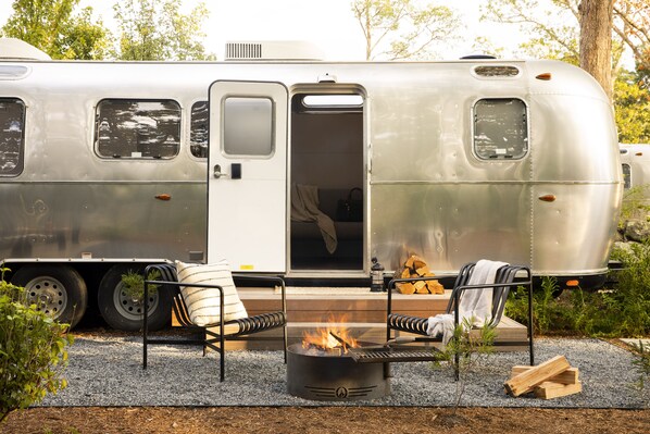 A stationary trailer with a fire pit and seating area, surrounded by trees and gravel.