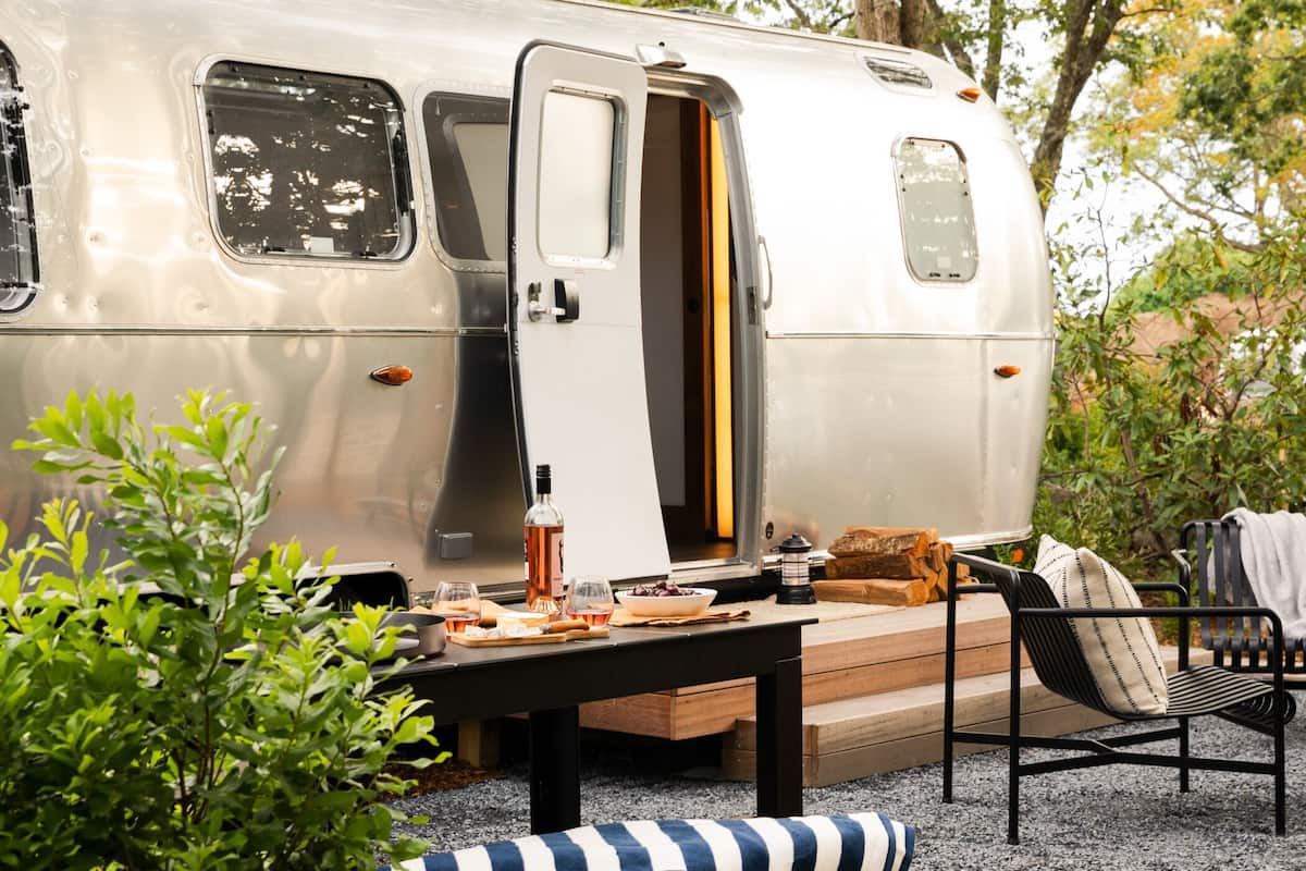 Una caravana plateada con terraza de madera, mobiliario exterior y una fogata.