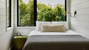 A bedroom with a bed, pillows, a bedside table with a clock and books, and a window with a view of trees. - AutoCamp Cape Cod (Falmouth)