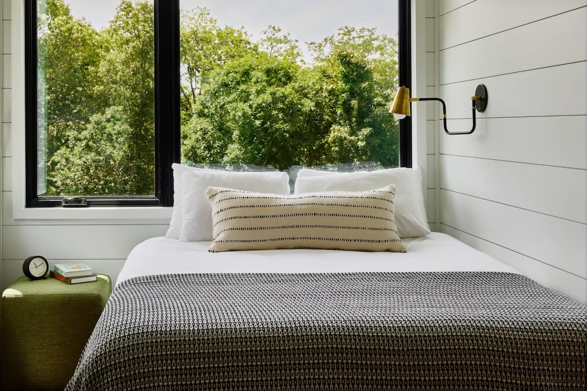 Un dormitorio con una cama, almohadas, una mesita de noche con un reloj y libros, y una ventana con vistas a árboles.