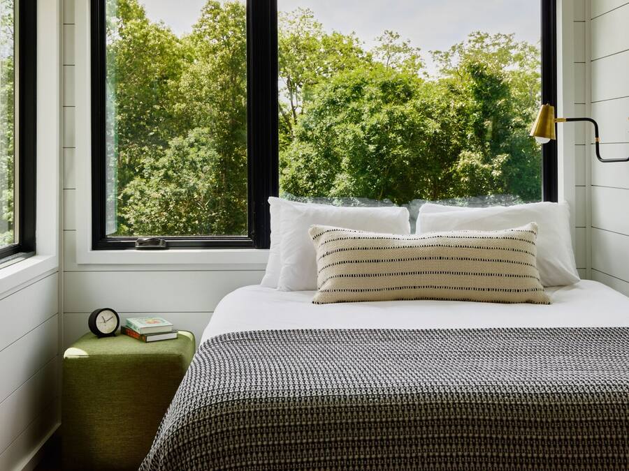 Une chambre à coucher avec un lit, des oreillers, une table de chevet avec une horloge et des livres, et une fenêtre donnant sur des arbres.