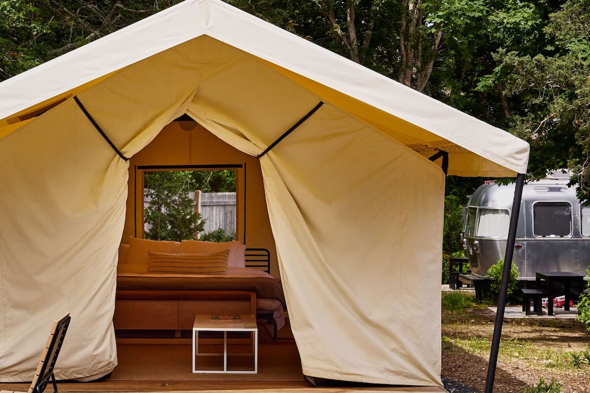 Una tienda de campaña con una cama en su interior, rodeada de árboles y una autocaravana estacionada.