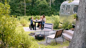 A group of people seated at an outdoor table with a grill, surrounded by lush greenery and a vintage camper.