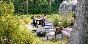 A group of people seated at an outdoor table with a grill, surrounded by lush greenery and a vintage camper.