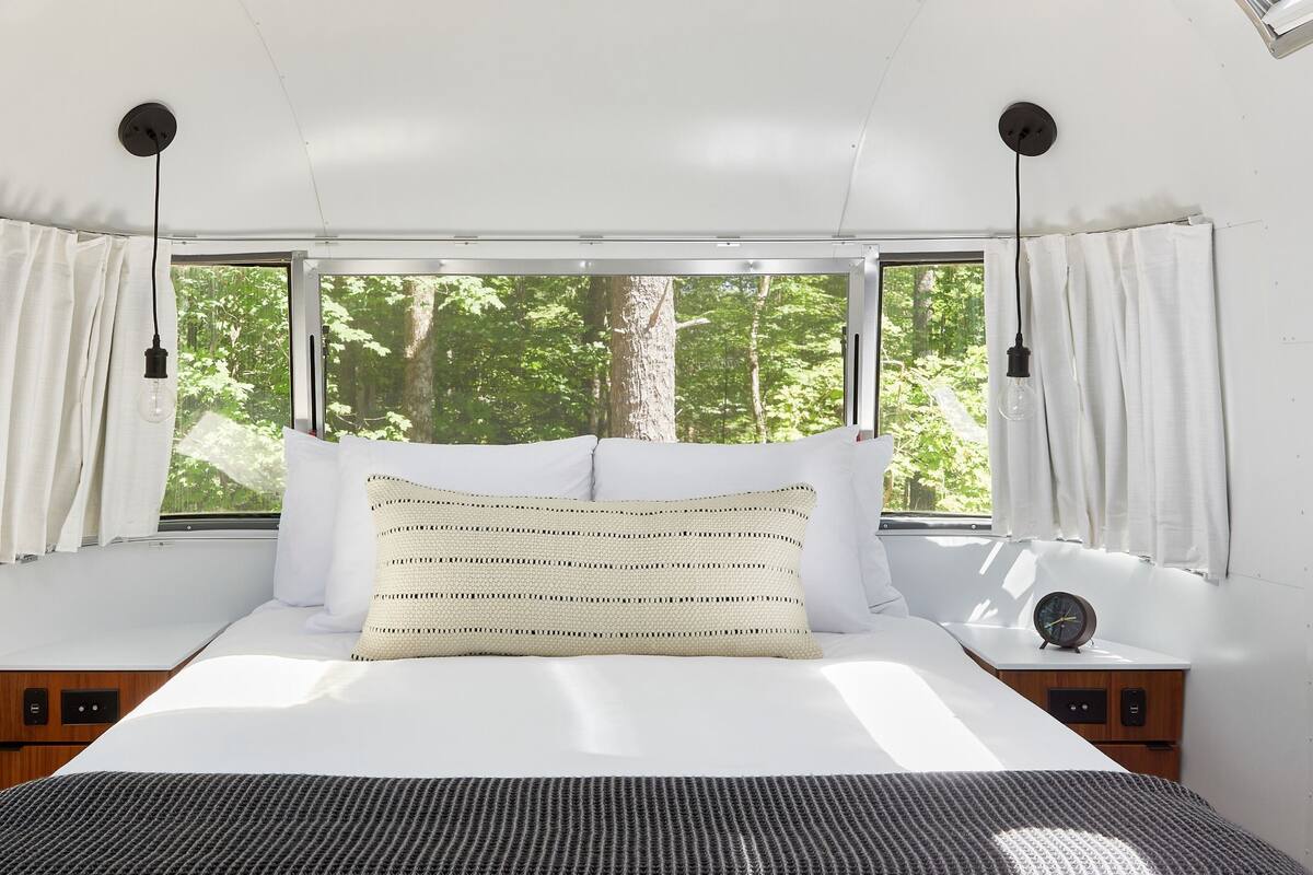 Una cama con un almohadón a rayas, una mesita de noche con un reloj y cortinas en una ventana con vistas a un bosque.