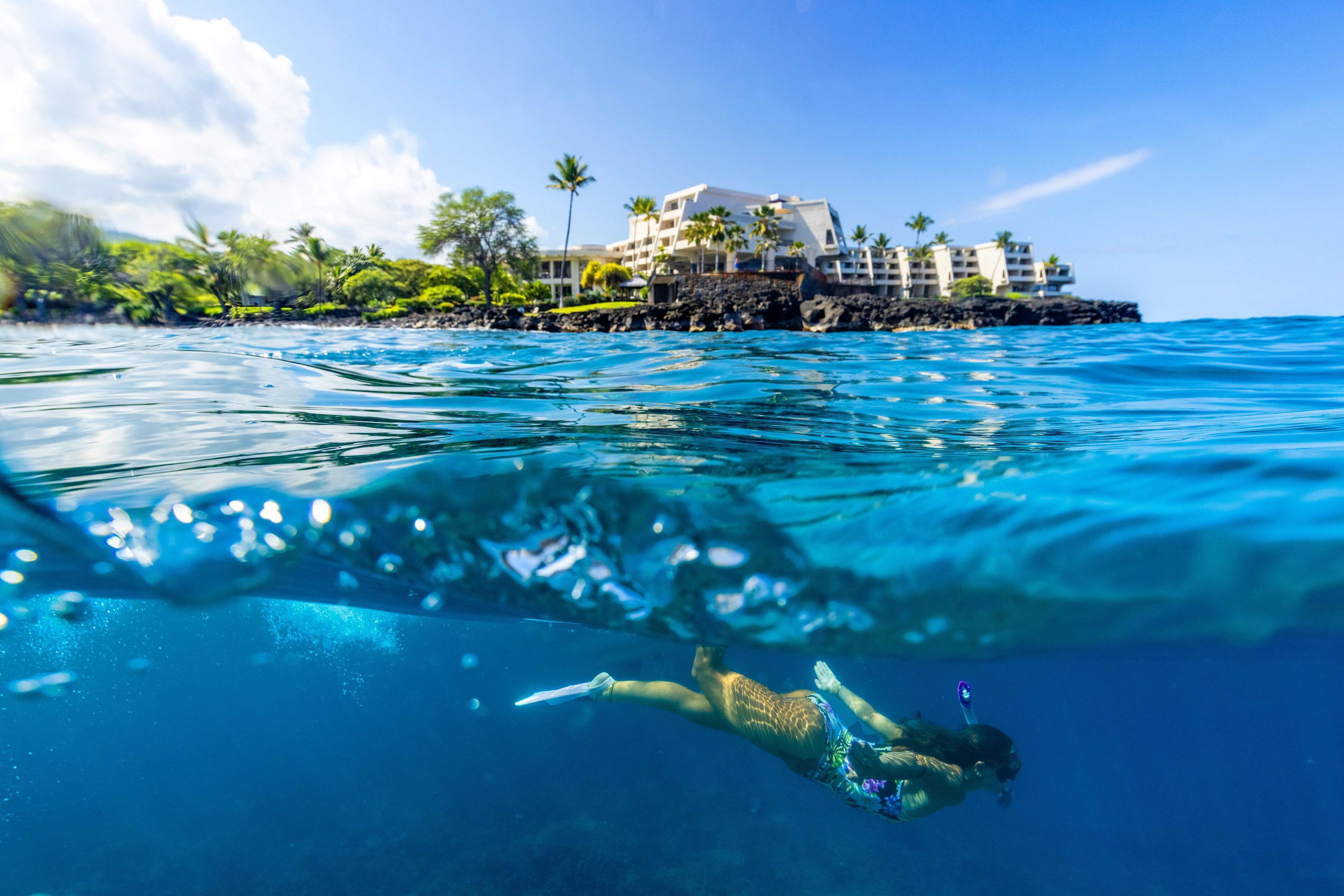 Un hôtel situé sur une île rocheuse, bordé d’eaux d’un bleu cristallin, un plongeur sous l’eau et un ciel dégagé.