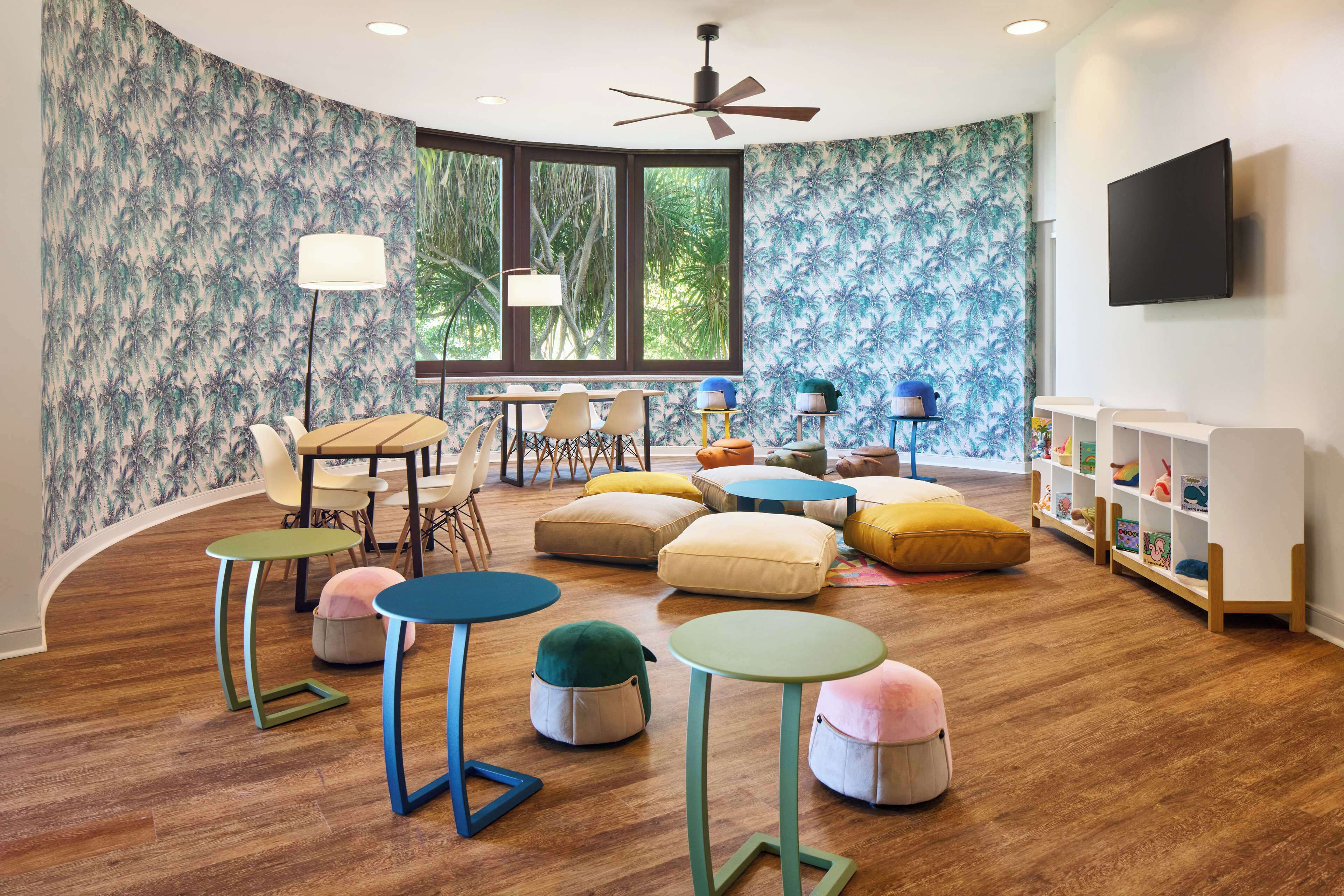 A room with a curved wall, wooden flooring, a ceiling fan, a TV mounted on the wall, a table with chairs, and a bookshelf.