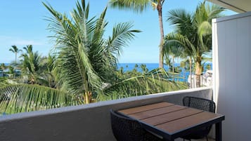 Varanda com mesa de madeira e cadeiras de vime, com vista para uma piscina e palmeiras, e uma visão clara do oceano.