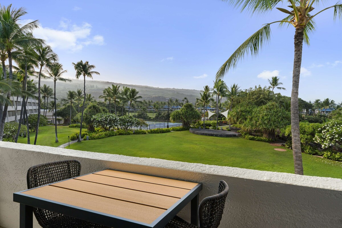 A balcony with a wooden table and wicker chairs, overlooking a well-maintained lawn with palm trees and a view of a distant mountain range.