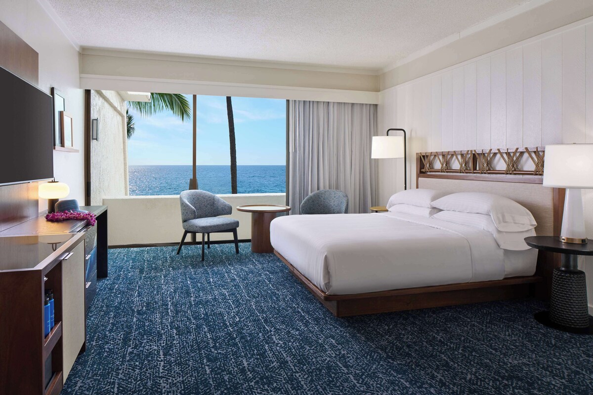 A hotel room with a bed, a desk, a chair, a TV, a window with a view of the ocean, and a lamp.