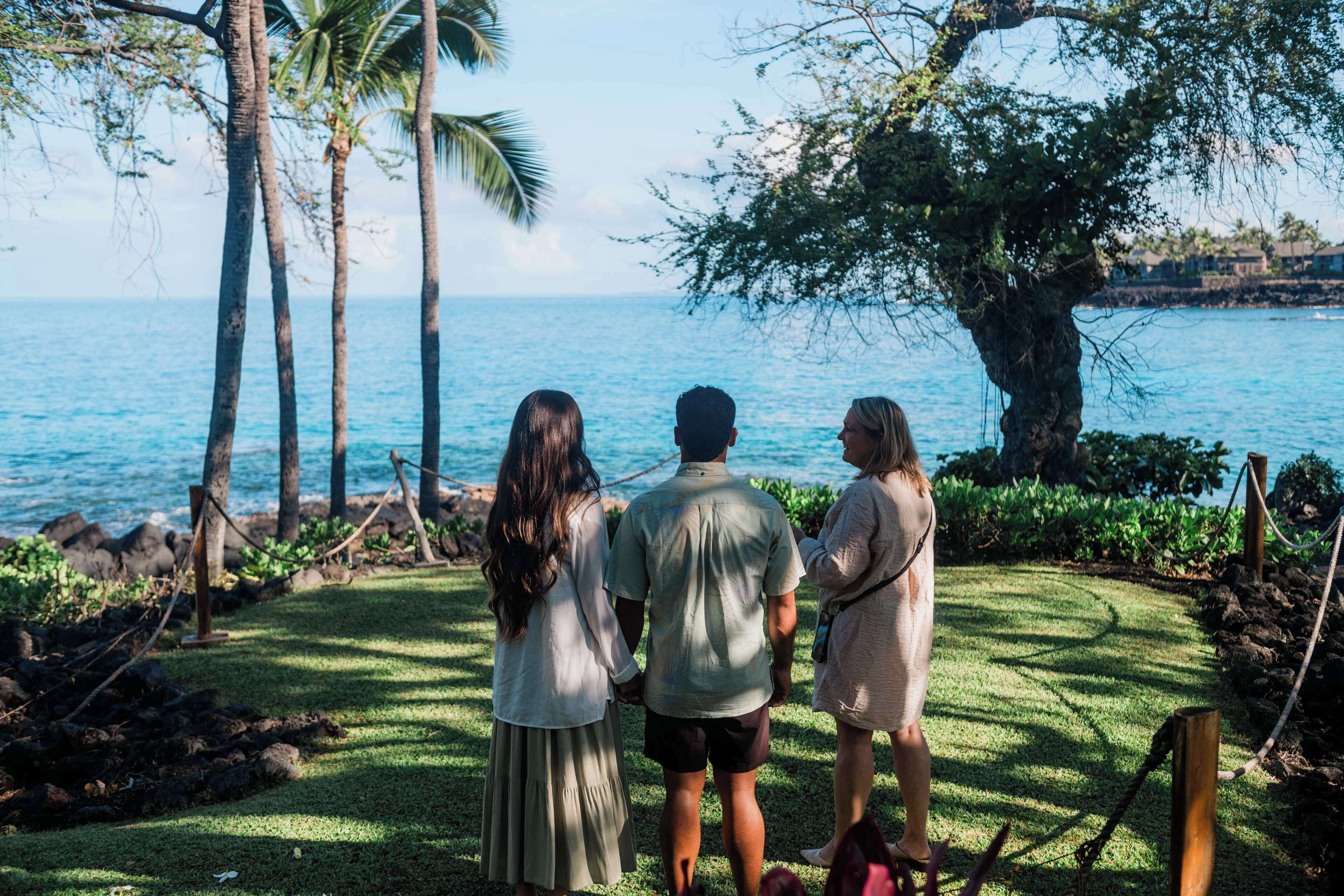 Three people standing on a grassy area with a view of the ocean and palm trees.