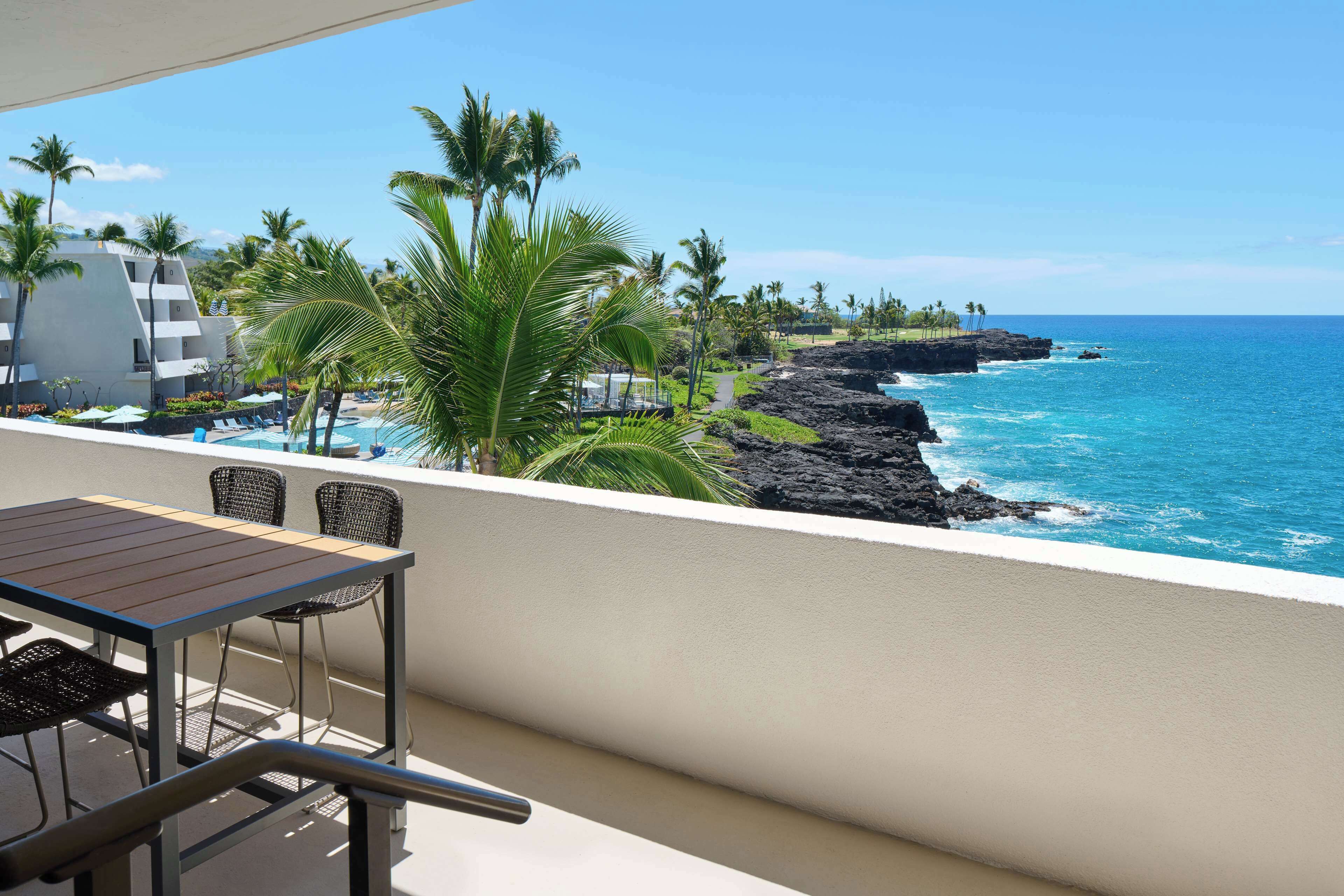 A balcony with a table and chairs, overlooking a pool and a coastline with palm trees and a lava rock formation.