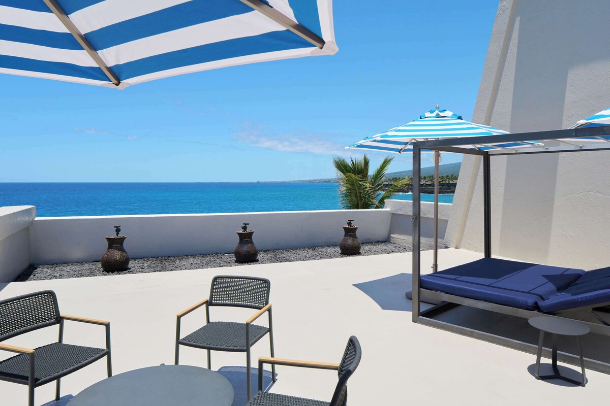 A terrace with a view of the sea, featuring a sunbed, two chairs, a table, and striped umbrellas.