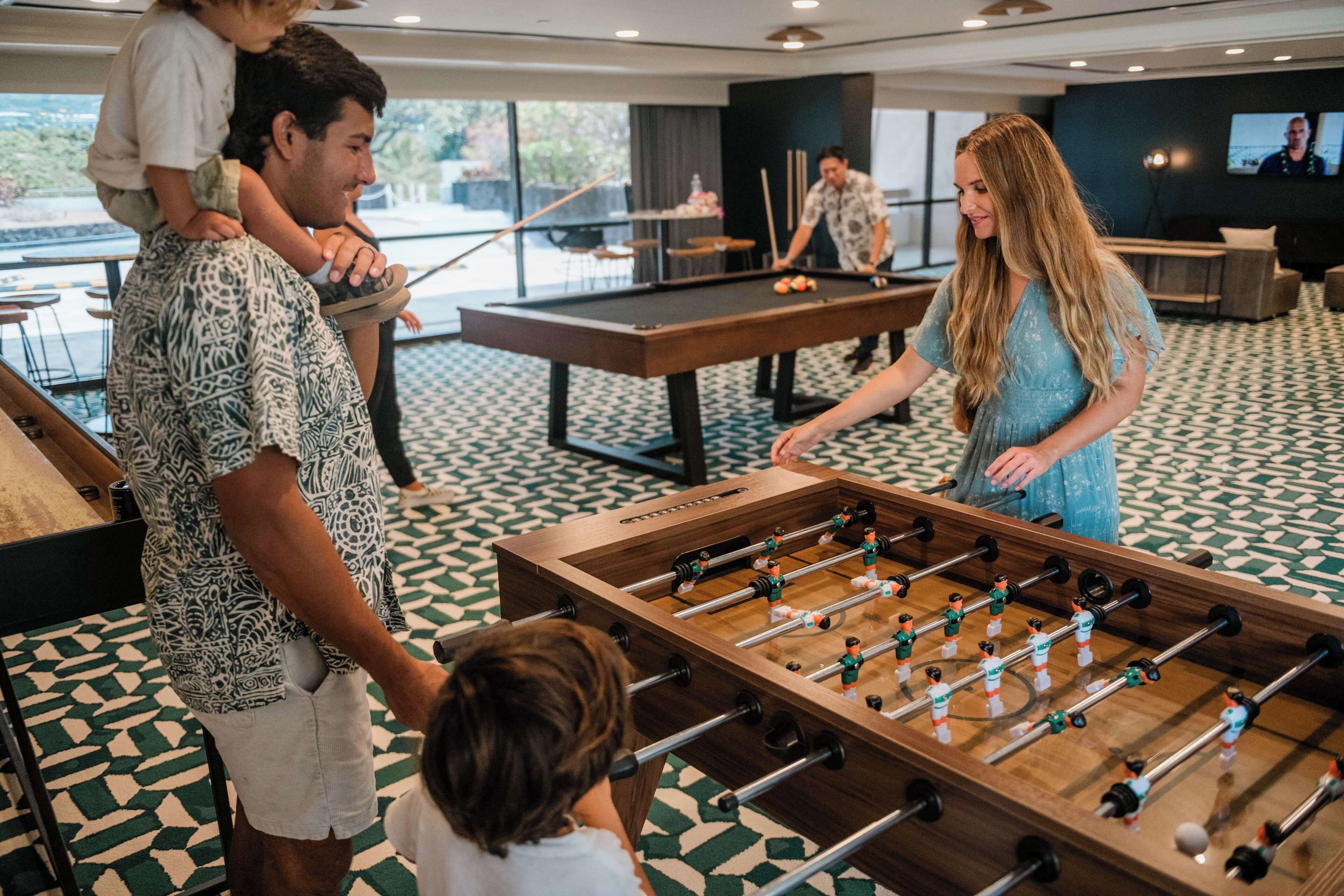 Mesa de billar y mesa de futbolín en el vestíbulo de un hotel, con piso estampado y ventanas grandes.