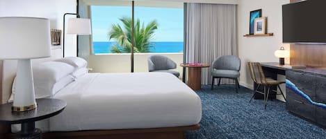 A hotel room with a bed, a desk, a chair, a TV, and a view of the ocean.