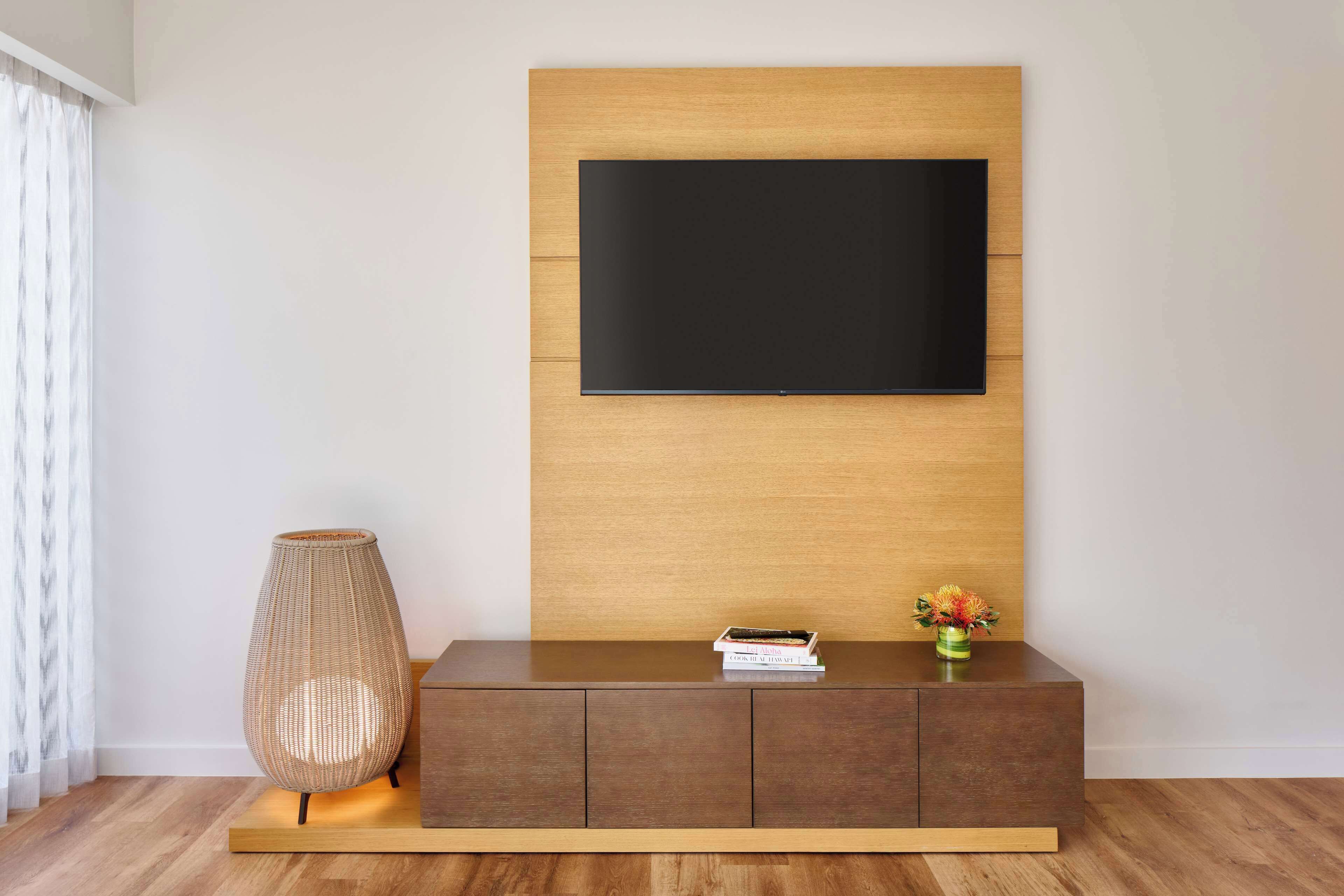 A modern living room with a flat-screen TV mounted on a wooden panel wall, a dark brown console table with a vase of flowers, and a tall woven lamp.