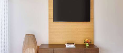 Una sala moderna con un televisor de pantalla plana montado en una pared de paneles de madera, una consola de mesa marrón oscuro con un florero y una lámpara alta de tejido.