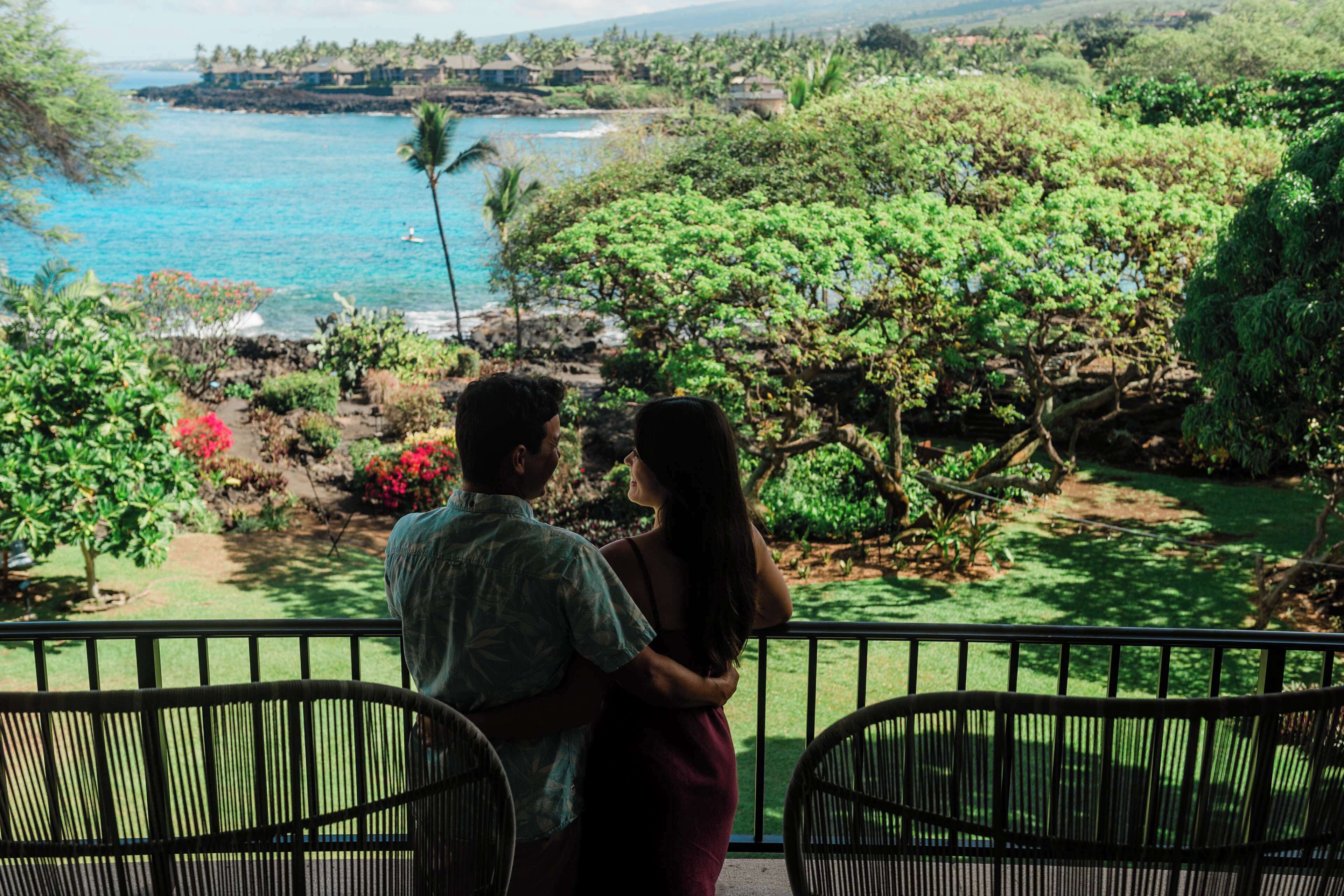 Una pareja en un balcón con vistas a un jardín exuberante y al mar.