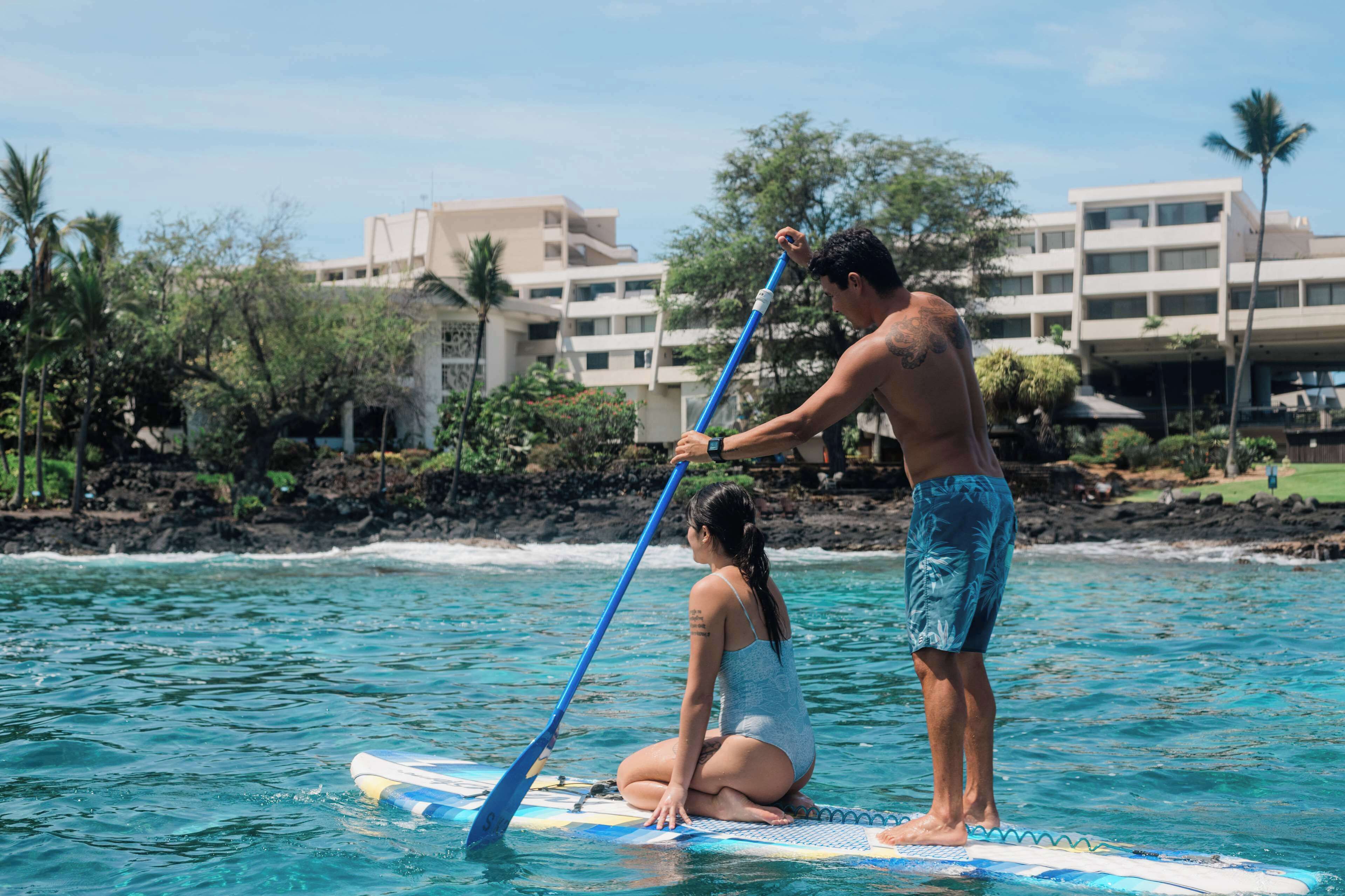 A man and a woman on a paddleboard in a coastal area with buildings in the background.