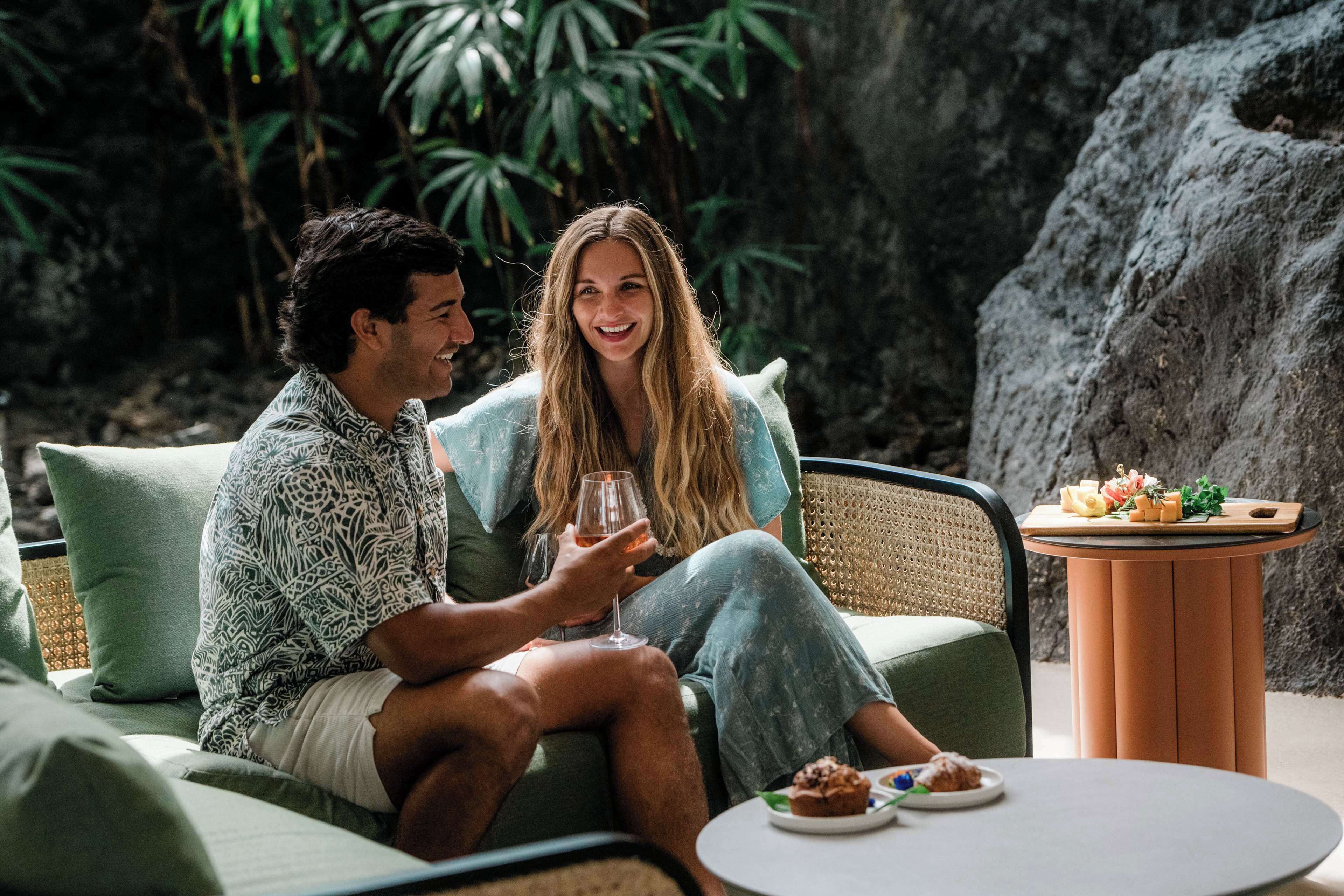 A couple sitting on a green sofa with a table between them, a small round table with food, and a rocky background.