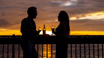 Silhuetas de um casal à mesa com bebidas, apreciando o pôr do sol sobre a água.