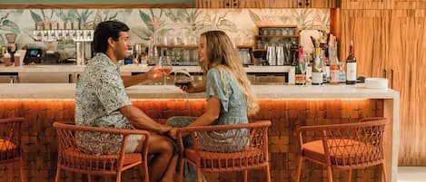 Un espace bar avec des fauteuils en osier, un comptoir équipé d’ustensiles de bar, et un décor mural composé d’un papier peint aux motifs tropicaux.